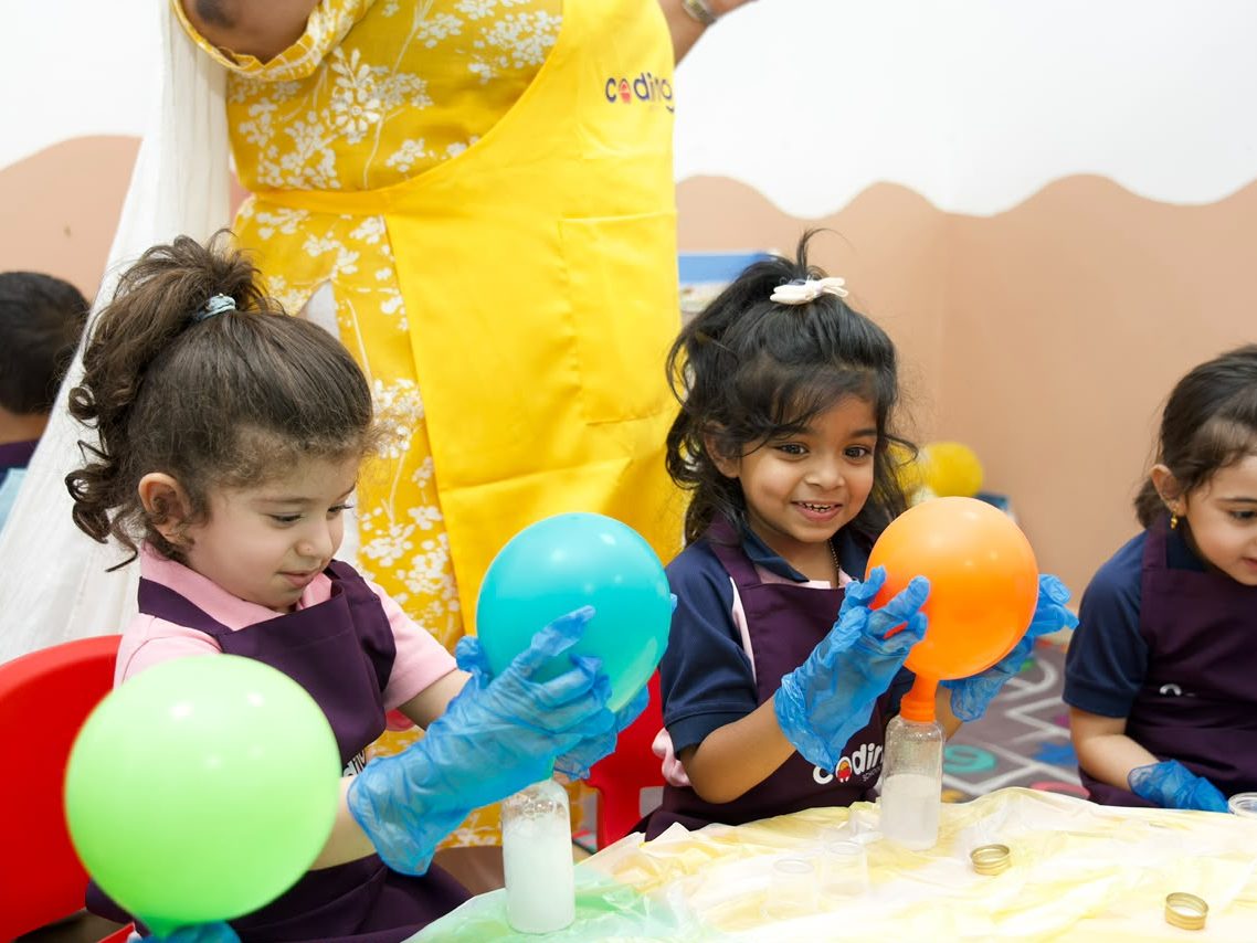 Nursery children in blue gloves holding green and orange balloons inflated by a vinegar and baking soda reaction.