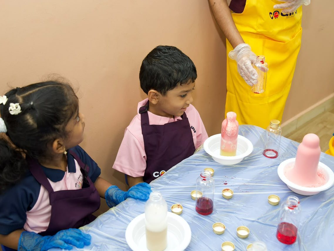 Two children in gloves at an experiment station watching a pink foam eruption rise from a bottle.