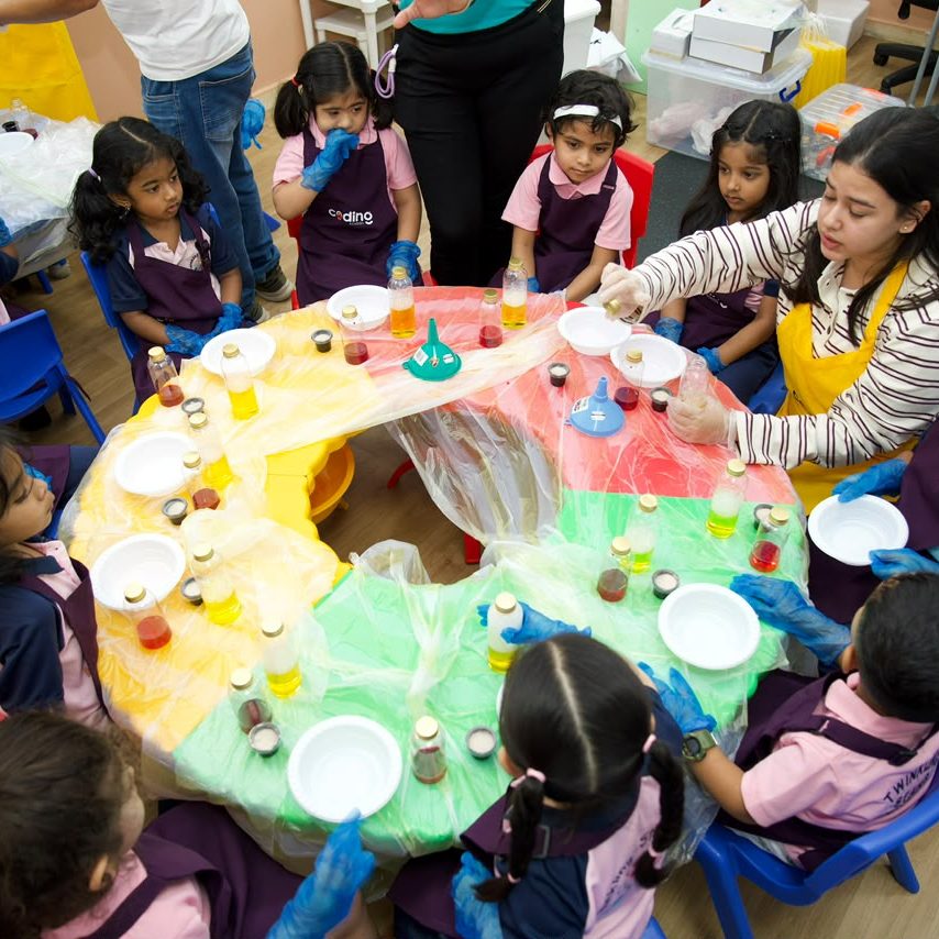 Children gathered around a colourful experiment table exploring materials with facilitators.