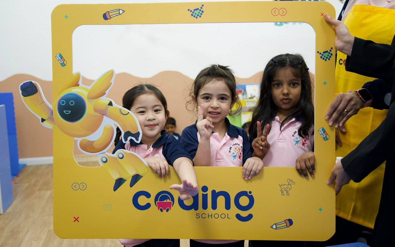 Three nursery children smiling through a bright yellow Coding School photo frame at a Tinker Fair.