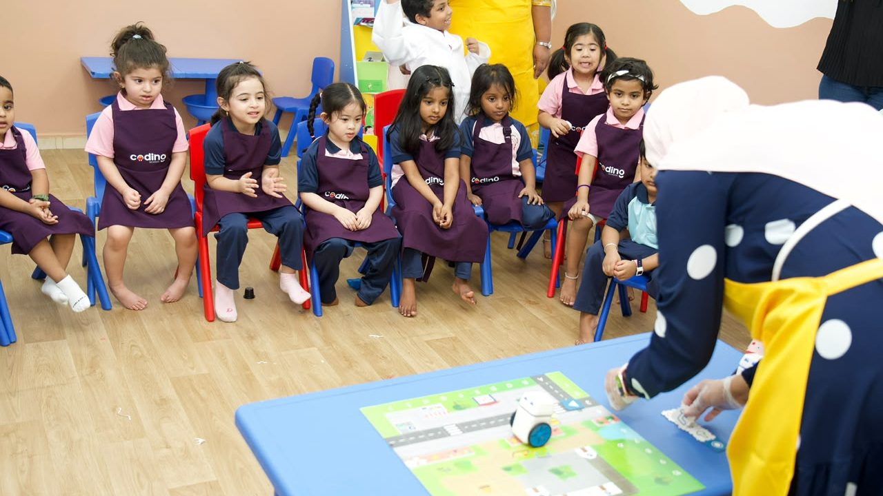 Children seated together watching the robotics zone demonstration on a blue table.