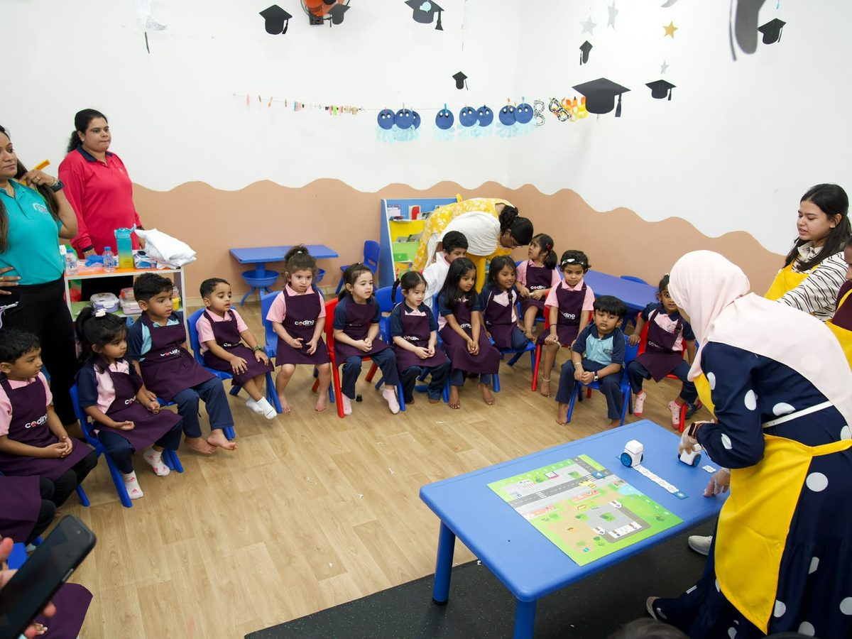 Nursery children seated in a session while a facilitator demonstrates a coding activity on a floor mat.
